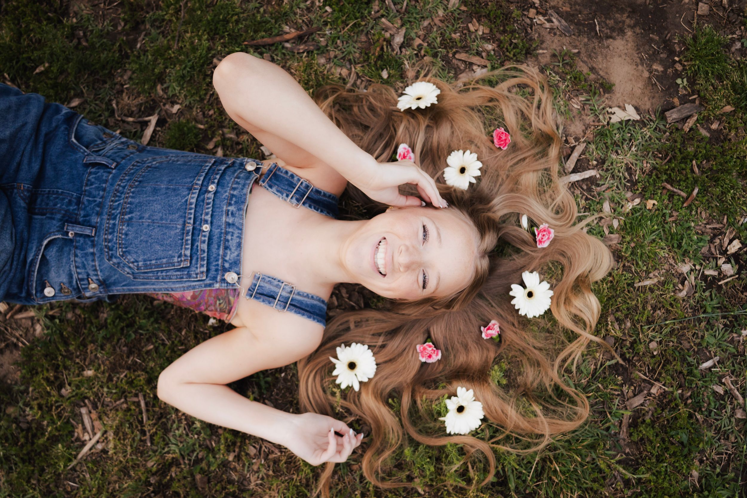 Joyful girl with daisy flowers in long wavy hair lying on grass in denim overalls.