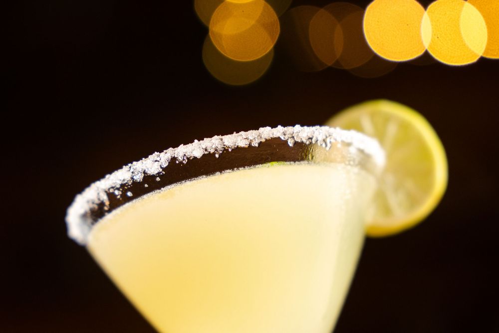 Chilled yellow margarita cocktail in salted rim glass garnished with lemon slice and bokeh lights.