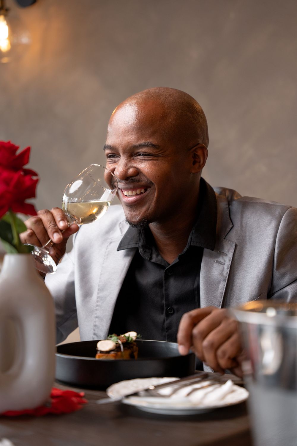 Happy bald man in silver blazer laughing while holding white wine glass at restaurant table.