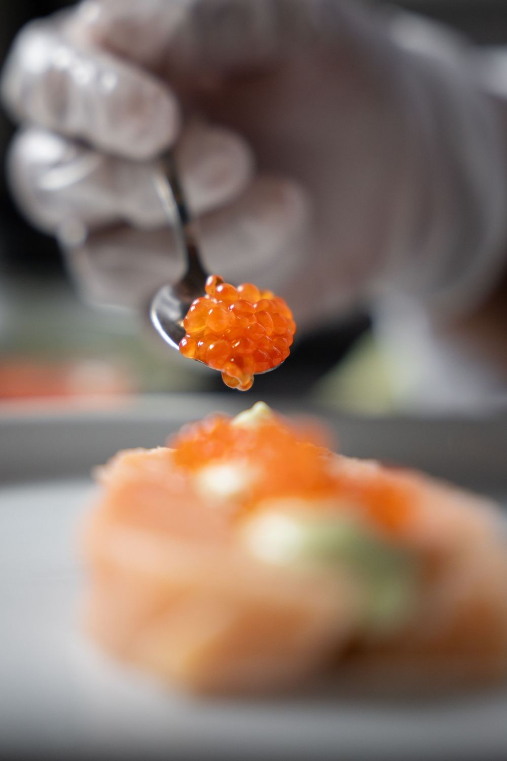 Chef in white glove placing orange salmon roe on top of fresh salmon dish with spoon.