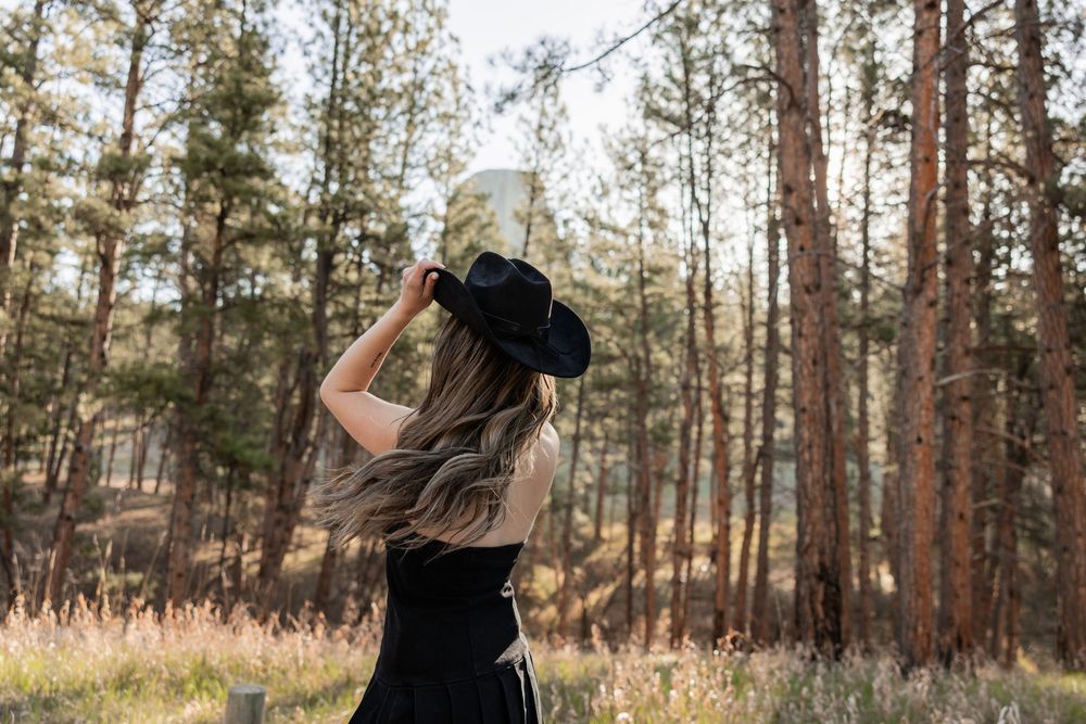 Woman in black dress and cowboy hat standing among tall pine trees in a sunlit forest clearing.