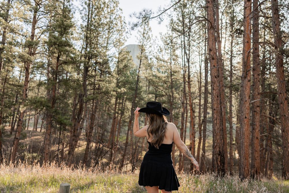 Woman in black dress and cowboy hat standing in a sunlit pine forest clearing.