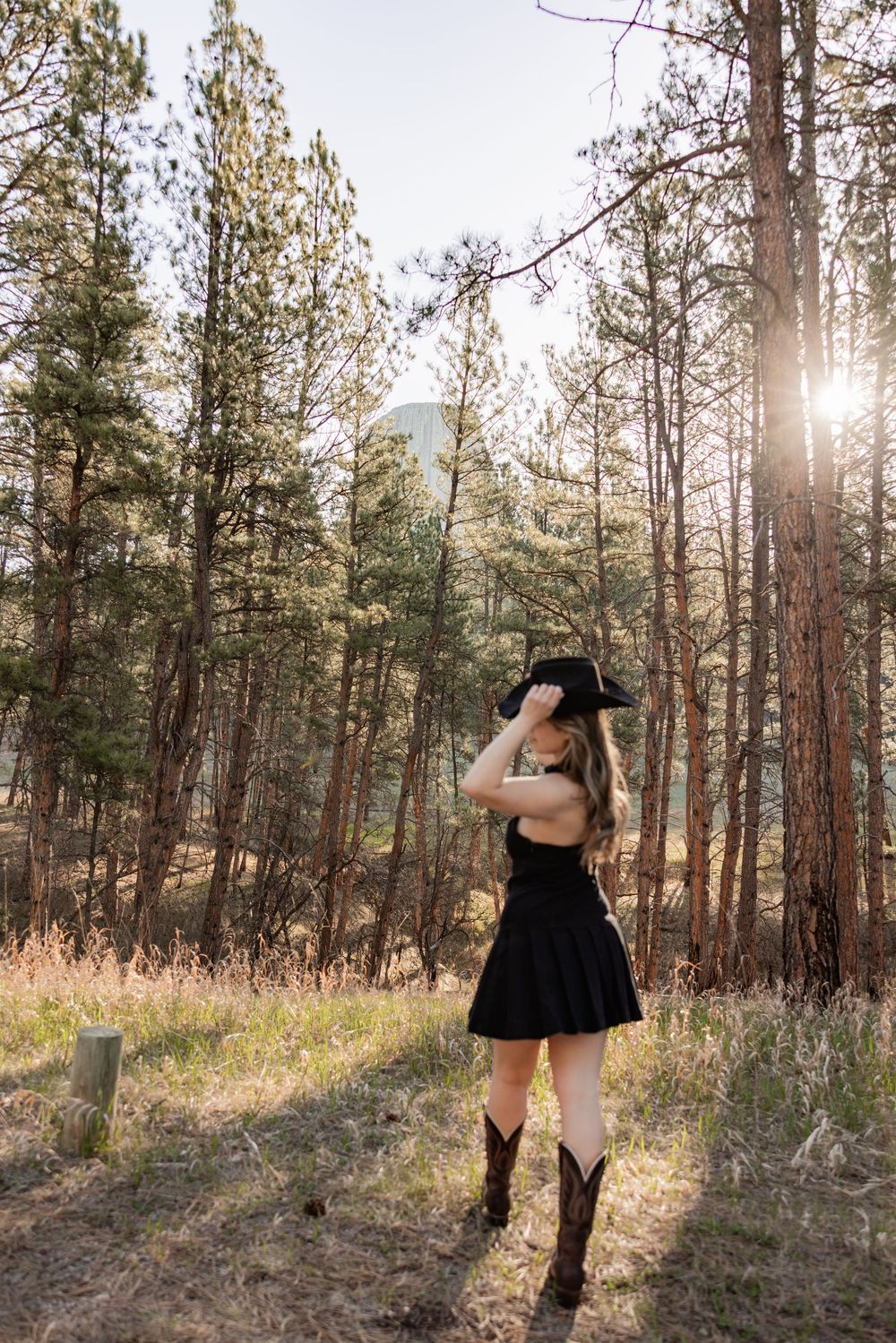 Woman in black dress and cowboy hat poses in sunlit pine forest wearing brown western boots.