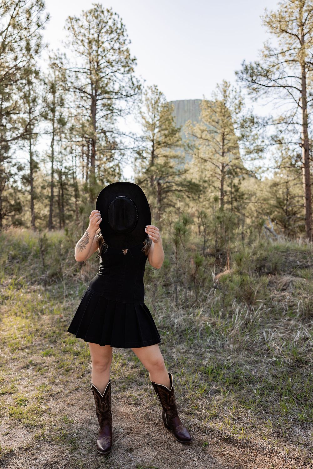 Woman in black dress and cowboy boots holds black hat over face in forest clearing with pine trees and rocky butte.