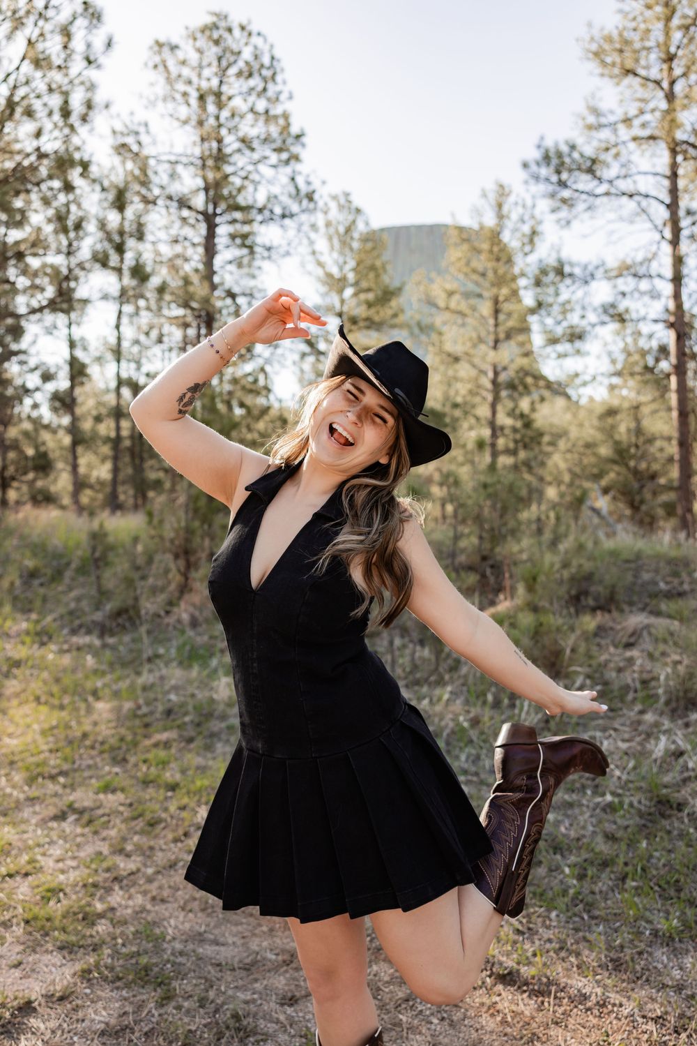 A joyful woman in a black dress and cowboy hat poses playfully in a pine forest, lifting one leg and smiling.