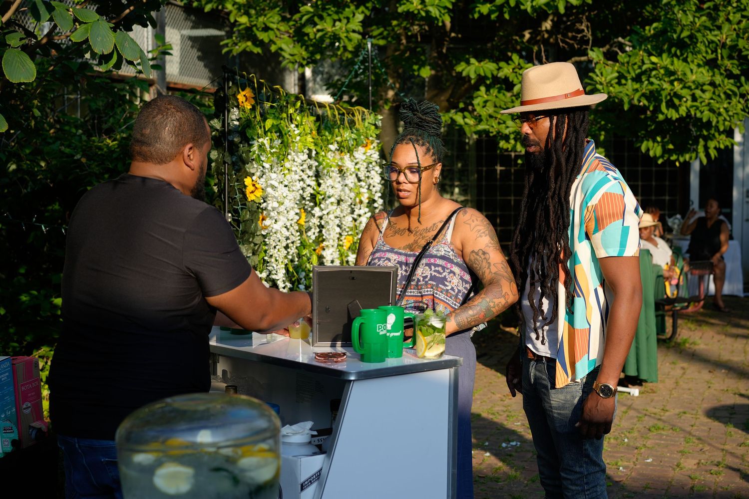 Three people interact at an outdoor vendor booth surrounded by lush greenery at a tropical event.