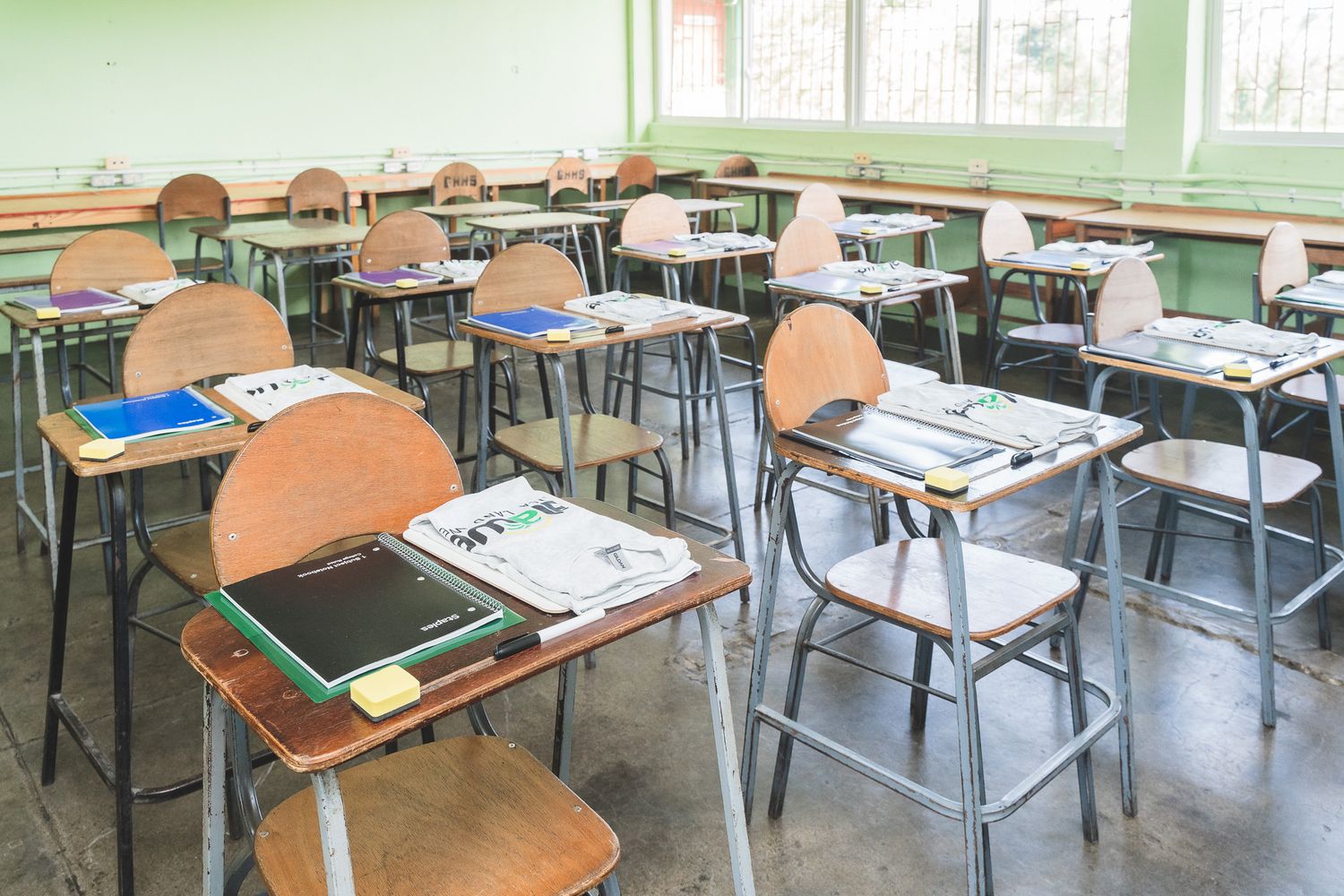 Empty classroom with wooden desks and chairs arranged in rows, books on desks, green walls, and large windows.
