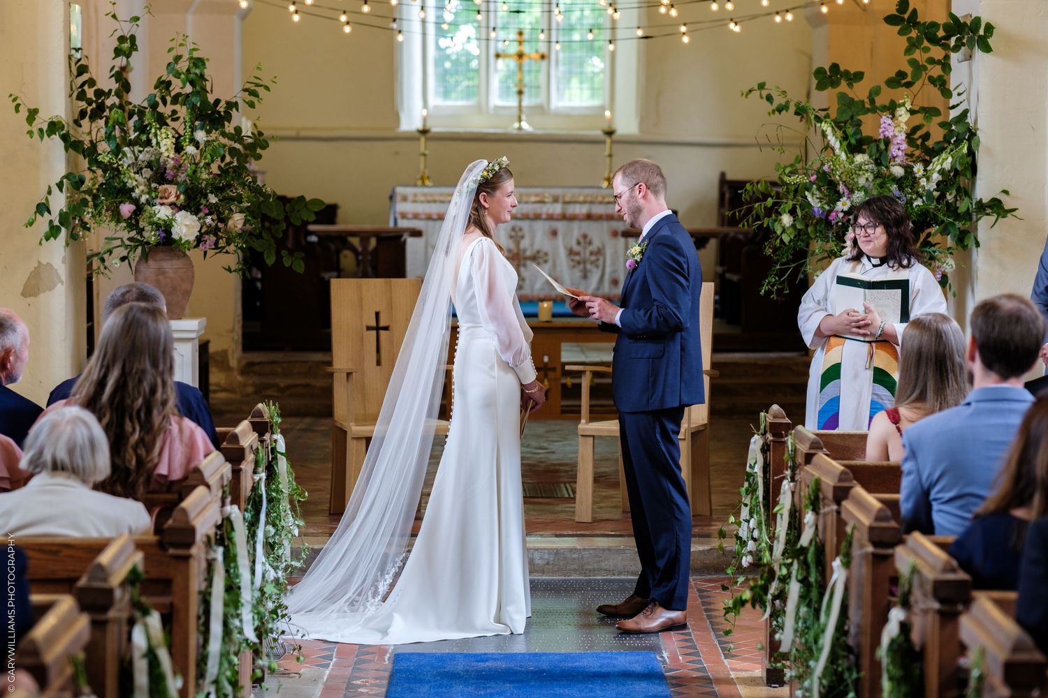 Bride and groom exchange vows at a floral-decorated church altar, officiated by a minister in a colorful stole.