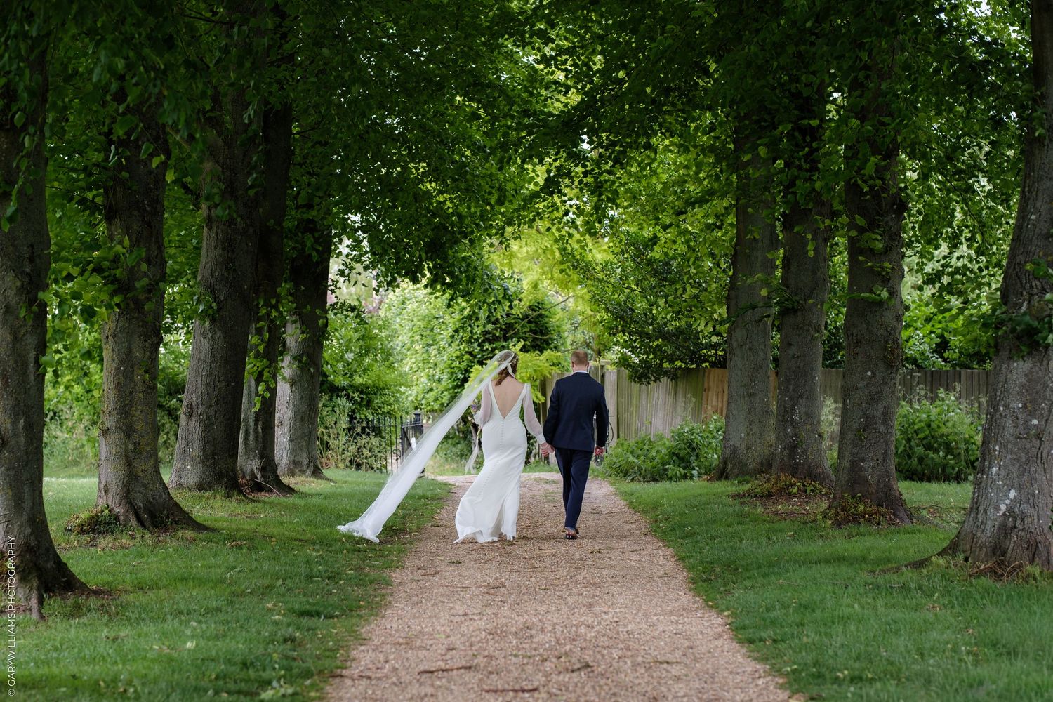 Bride and groom walk hand-in-hand down a tree-lined path, bride's veil flowing, in a lush green garden setting.
