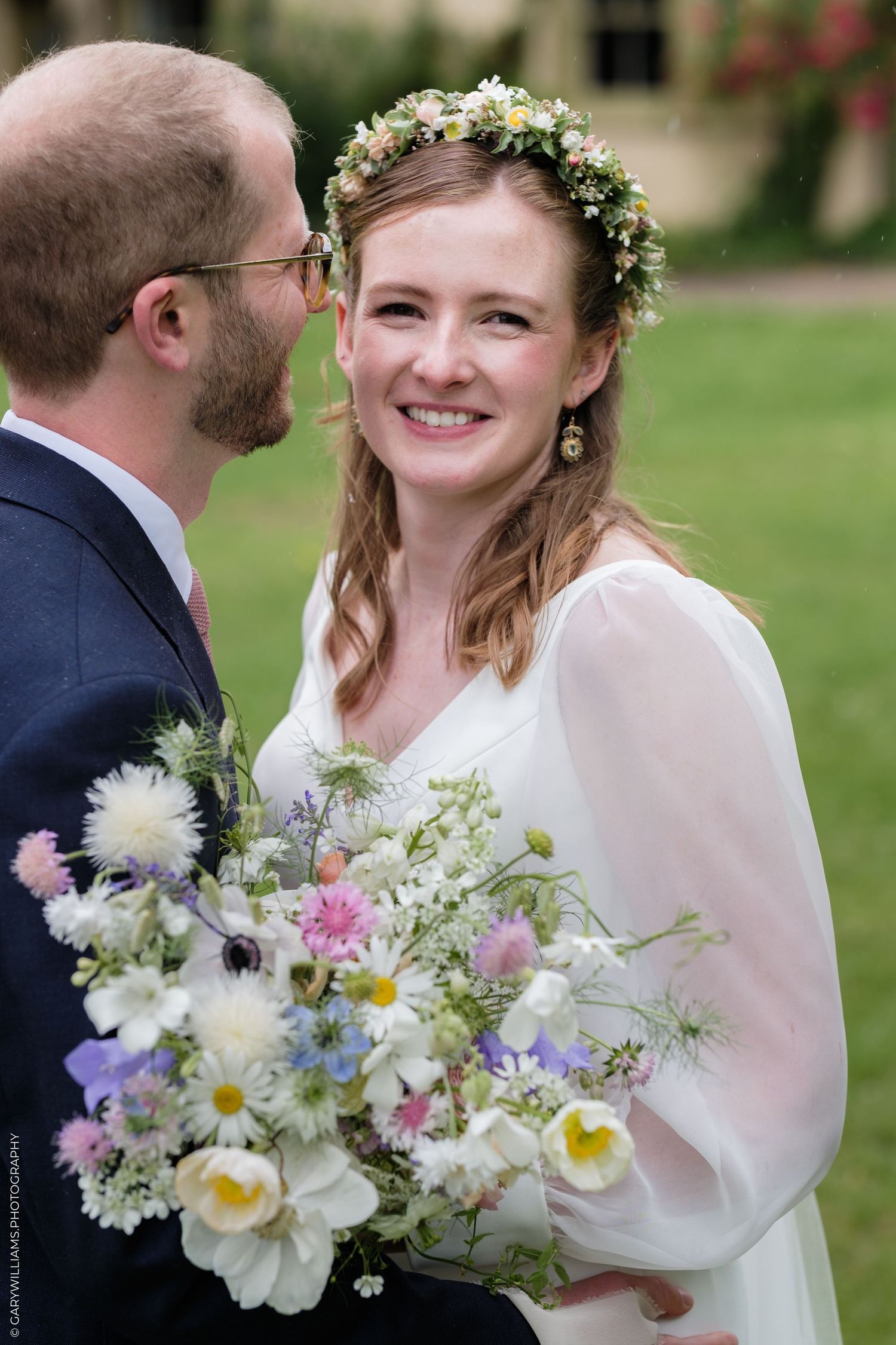 Smiling bride wearing floral crown holds wildflower bouquet with pastel blooms beside groom in navy suit.