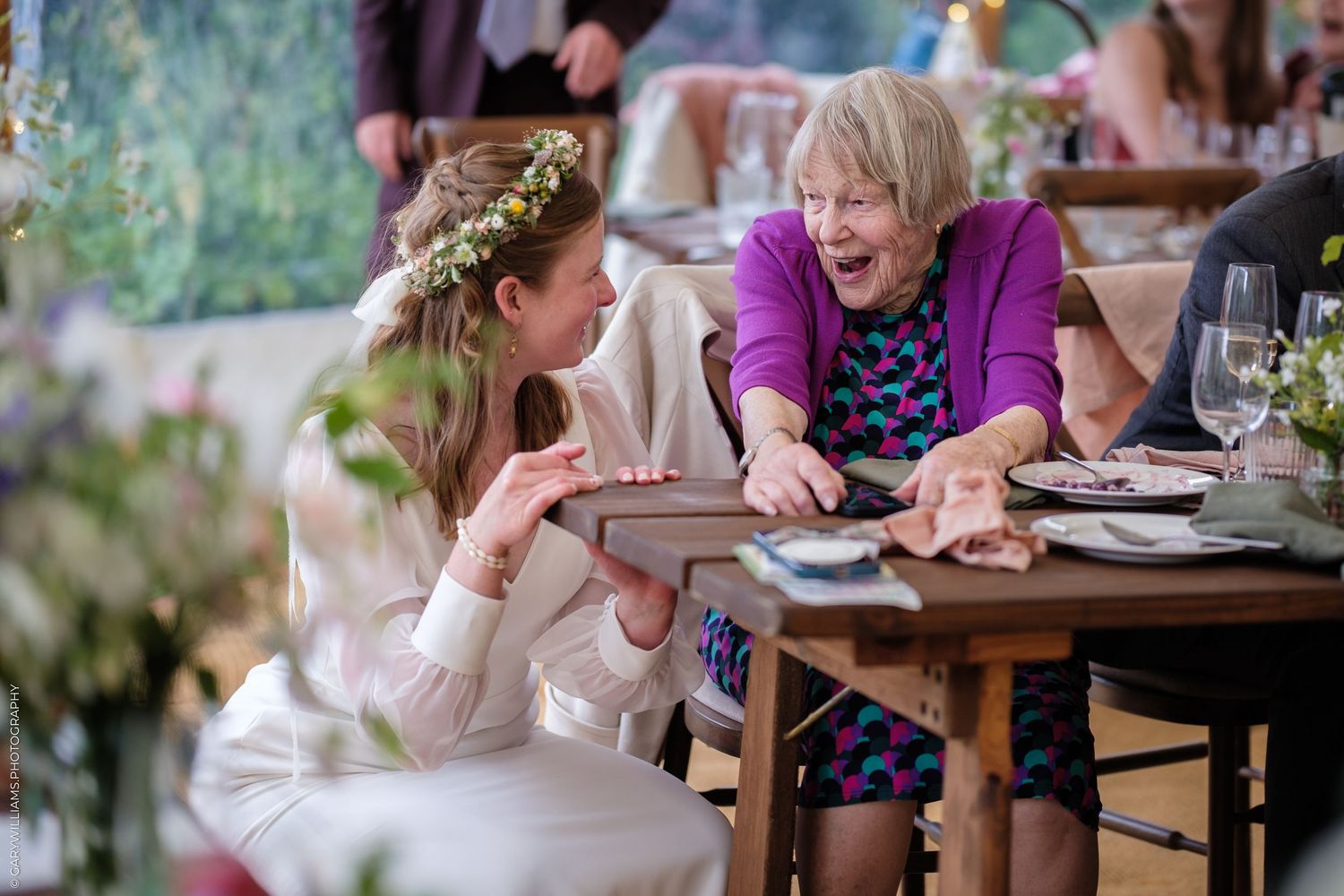 Bride in floral crown laughing joyfully with elderly woman in purple at wedding reception table.