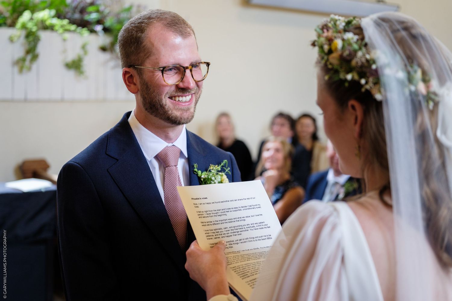 Smiling groom in navy suit reads wedding vows with bride in floral crown during intimate ceremony.