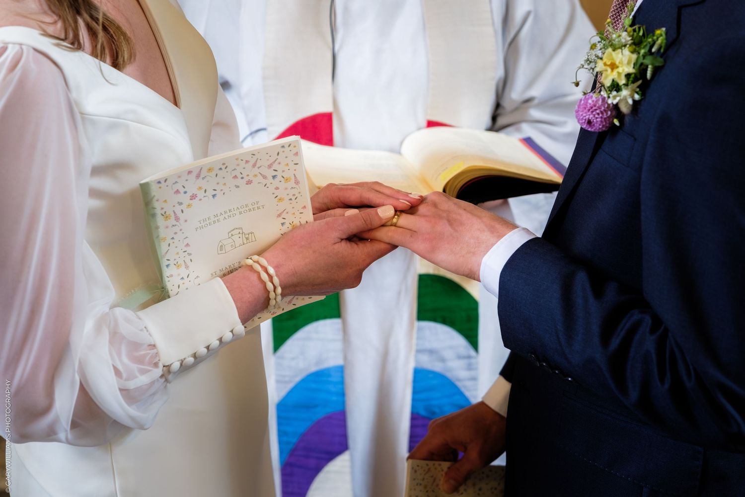 Bride and groom exchanging wedding rings during a church ceremony, officiant holding open book in background.