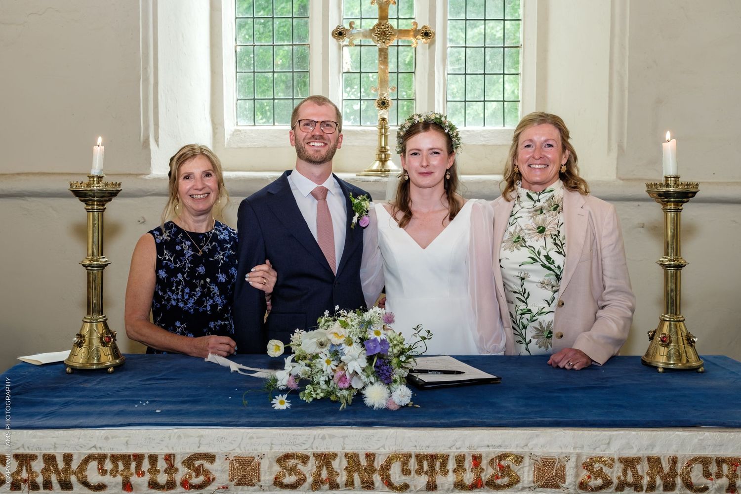 Bride and groom sign wedding register at church altar flanked by two women guests, with floral bouquet and candles.