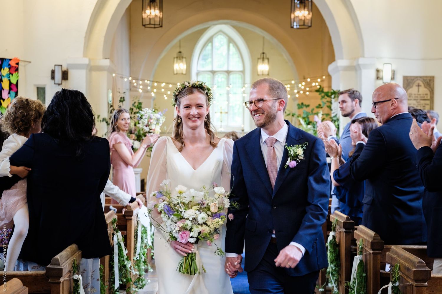 Bride and groom walk down church aisle after wedding ceremony, bride holding wildflower bouquet wearing floral crown.