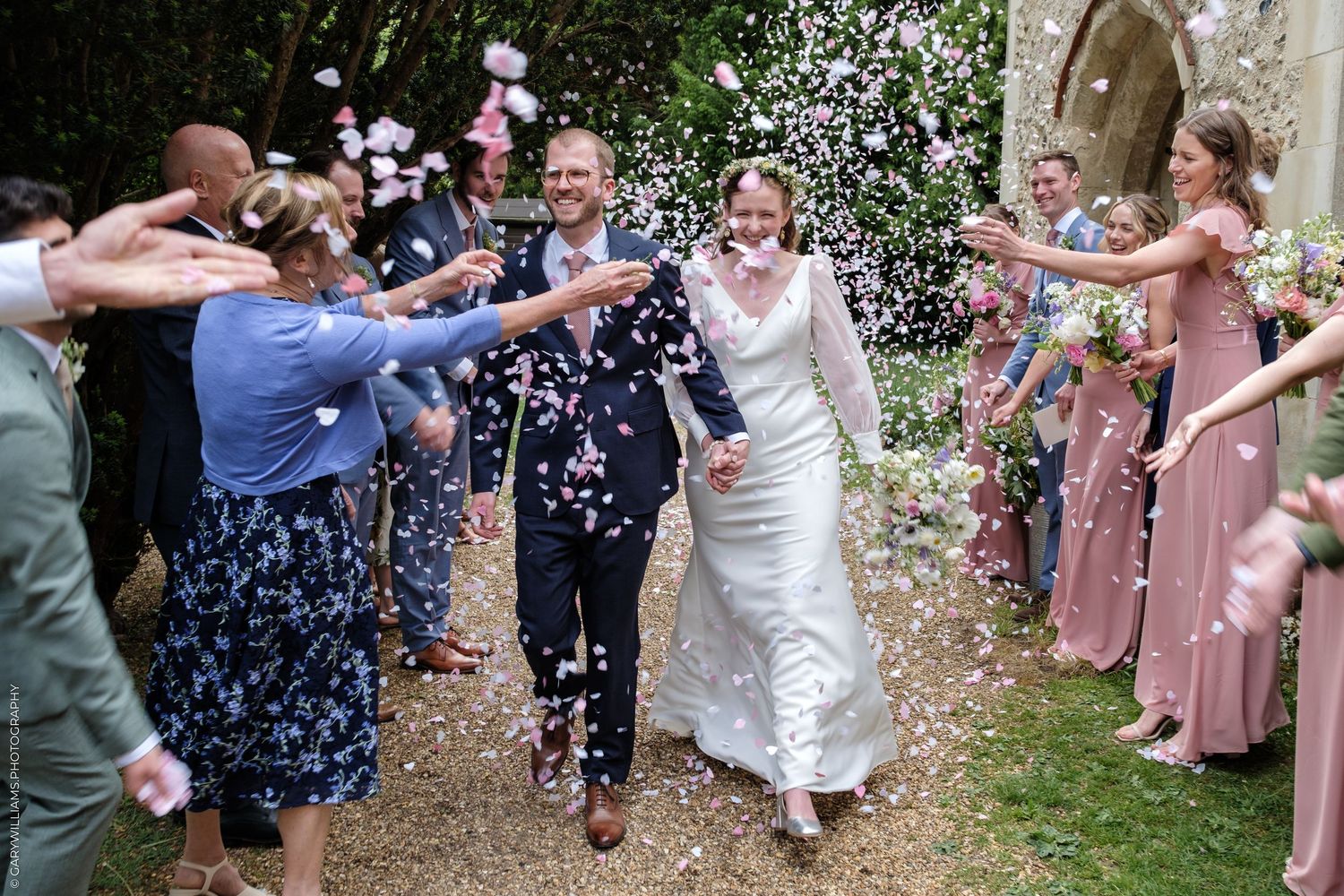 Newlyweds walk through confetti shower as guests celebrate outside historic stone church after wedding.