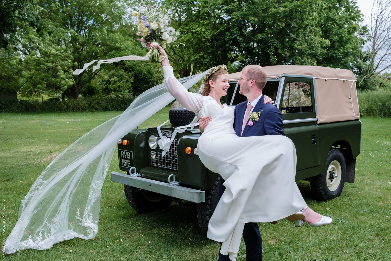Bride and groom posing joyfully with vintage green Land Rover on grass, veil flowing in the breeze.