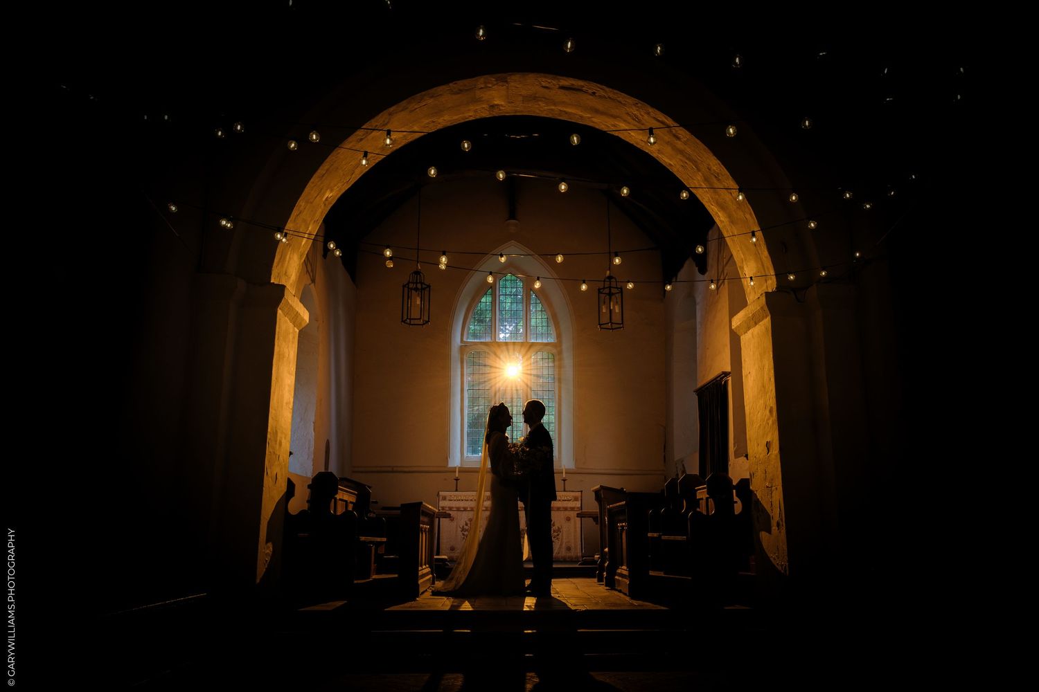 Silhouette of a couple standing under a glowing arch in a dimly lit chapel with string lights and stained glass window.