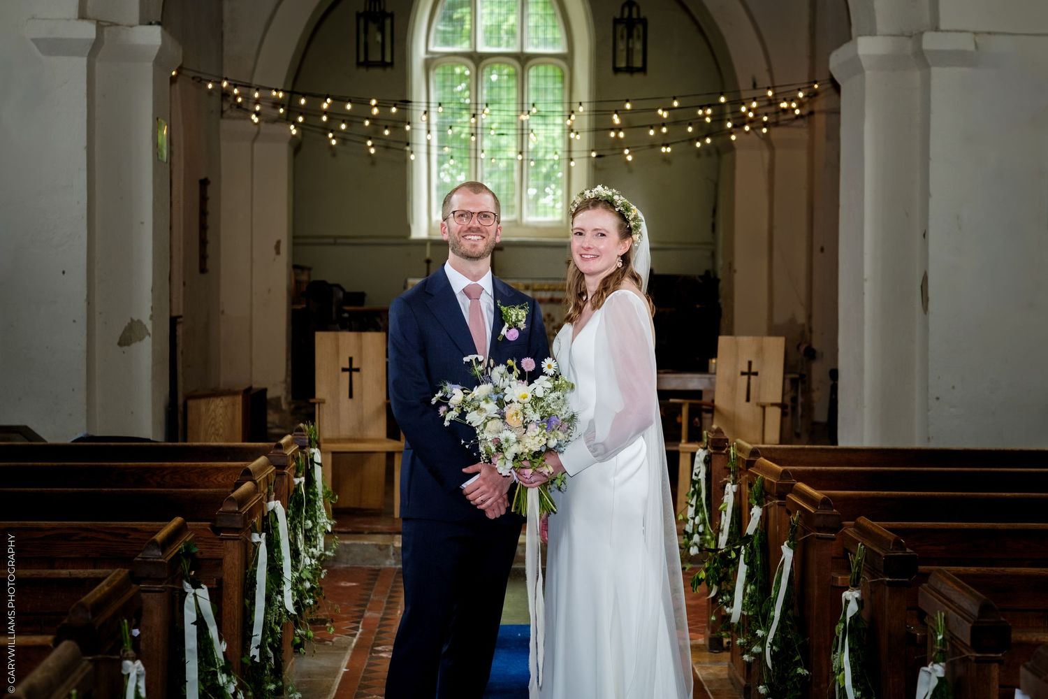 Bride and groom smiling together inside a decorated church, bride holding a white floral bouquet on their wedding day.