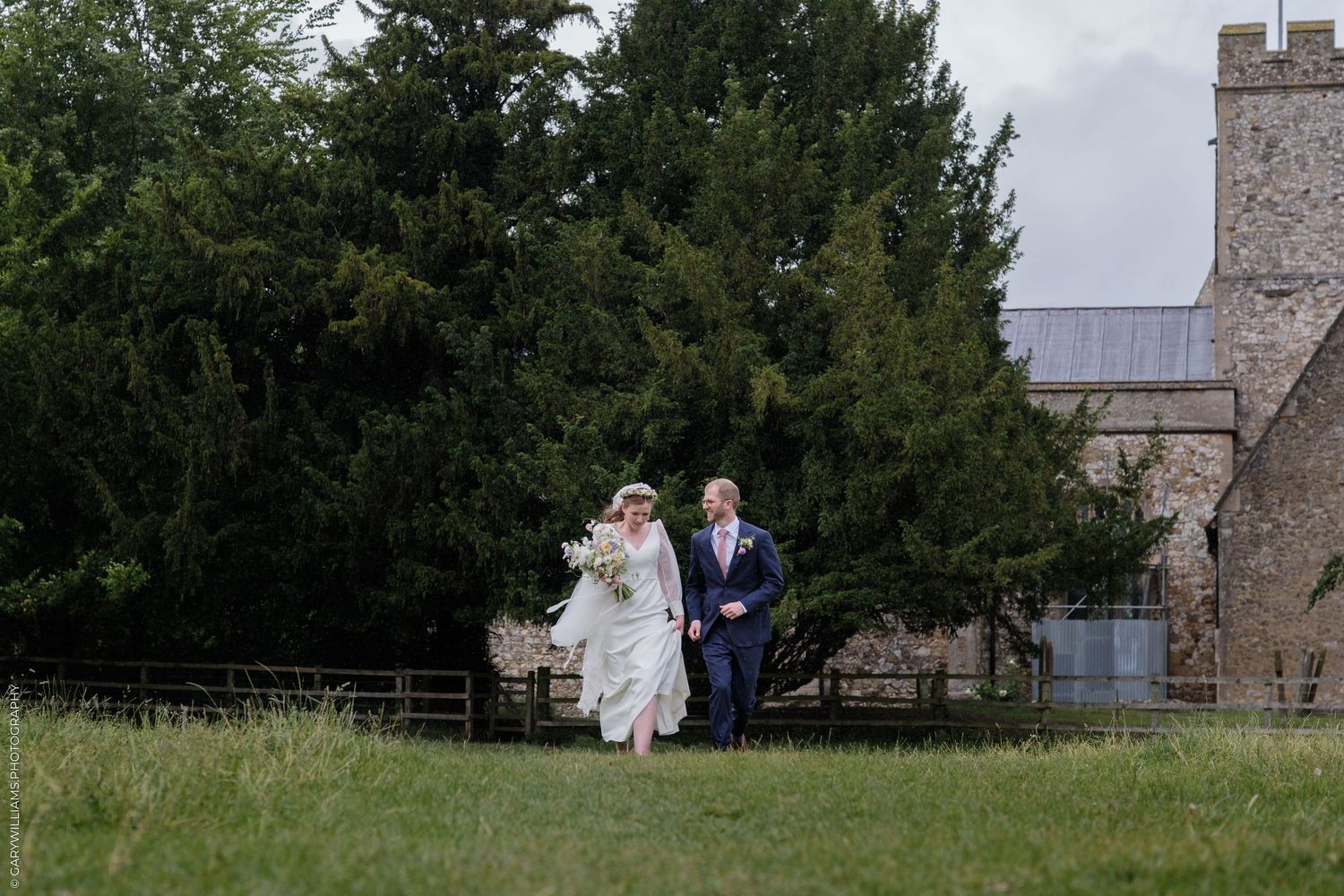 Bride and groom walking together on green lawn beside stone church surrounded by tall trees on their wedding day.