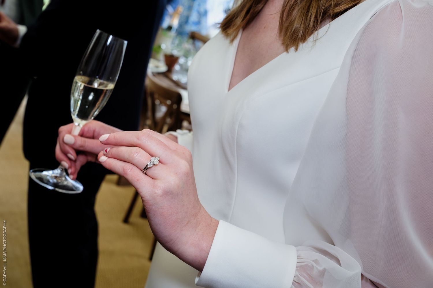 Bride in white dress holding champagne flute, showcasing diamond engagement ring at wedding reception.