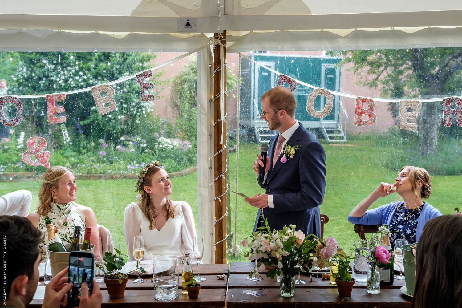 Groom giving a wedding speech at a decorated marquee reception while bride and guests listen attentively.