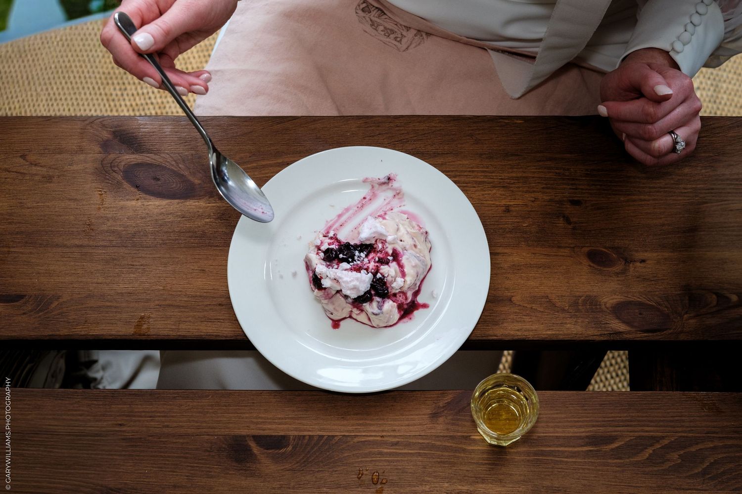 Overhead view of a dessert plate with berry cream on a wooden table, with a person holding a spoon nearby.