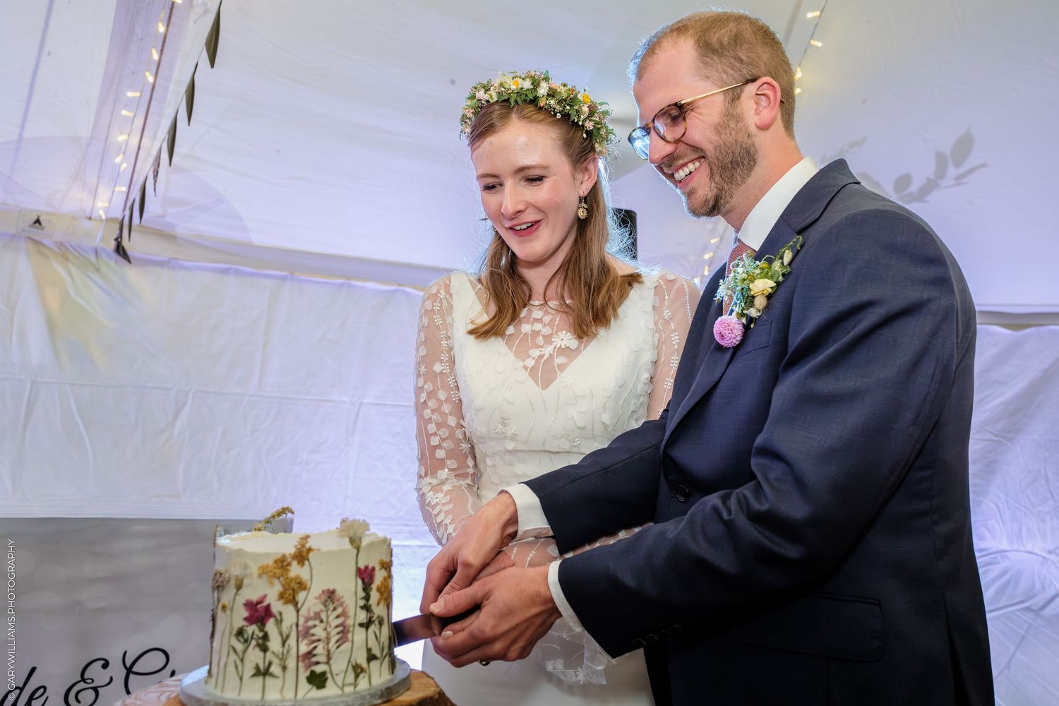 Bride and groom cutting their floral wedding cake, bride wearing flower crown and lace dress, groom in navy suit.