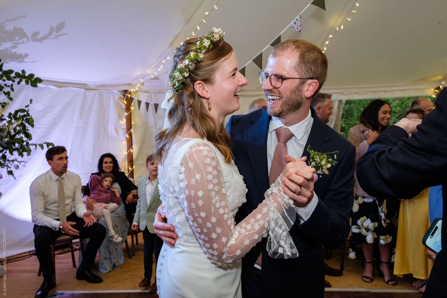 Bride and groom share first dance in decorated wedding marquee, bride wearing floral crown and white lace dress.