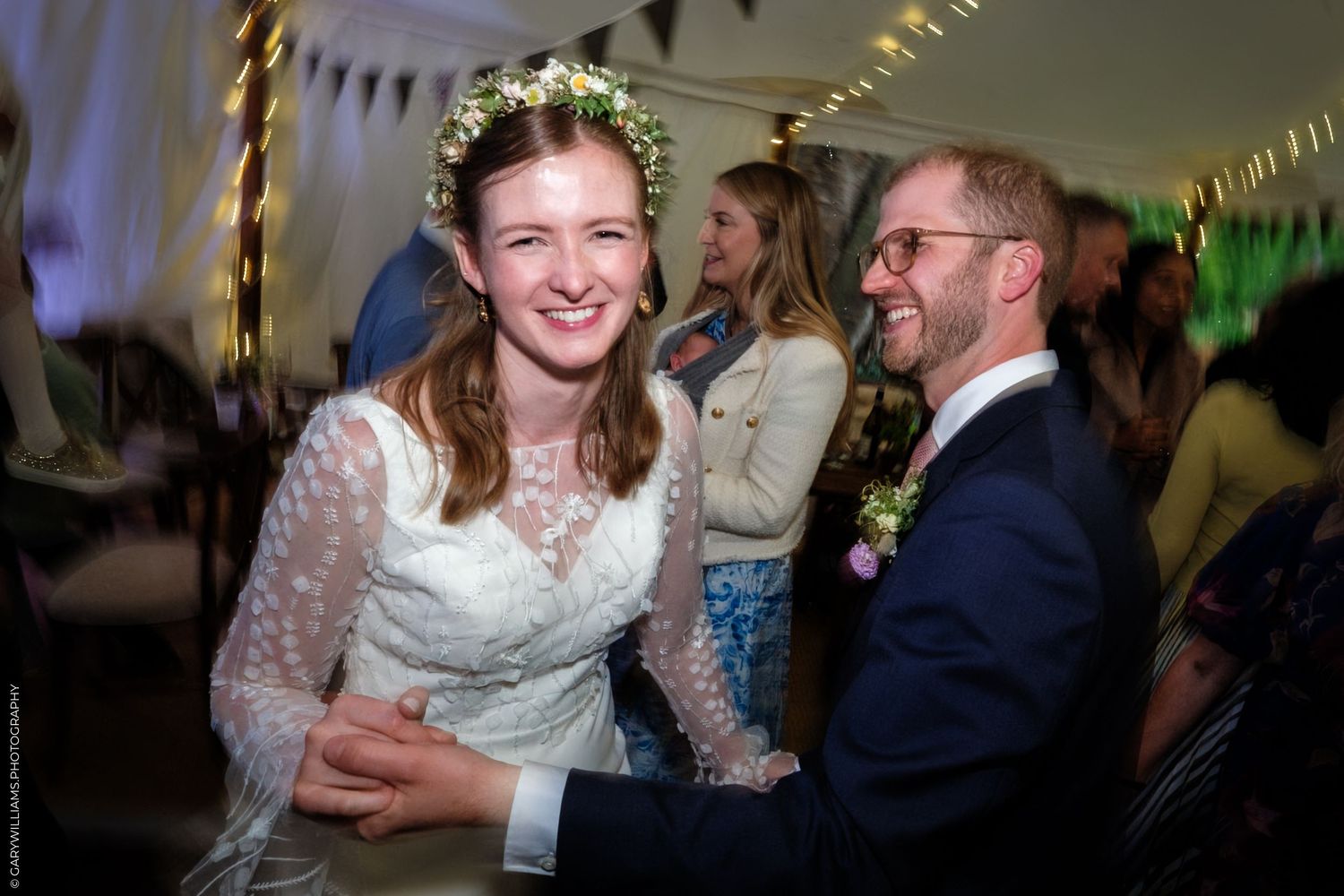 Bride in white lace dress and flower crown laughing with groom in navy suit at wedding reception.
