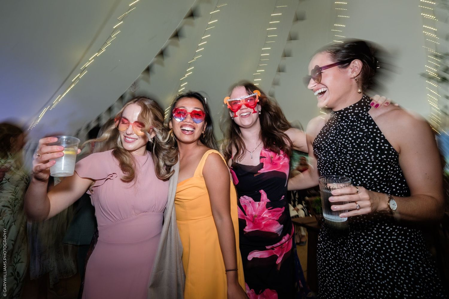 Four women in colorful dresses and fun sunglasses laughing and holding drinks at a festive wedding reception party.