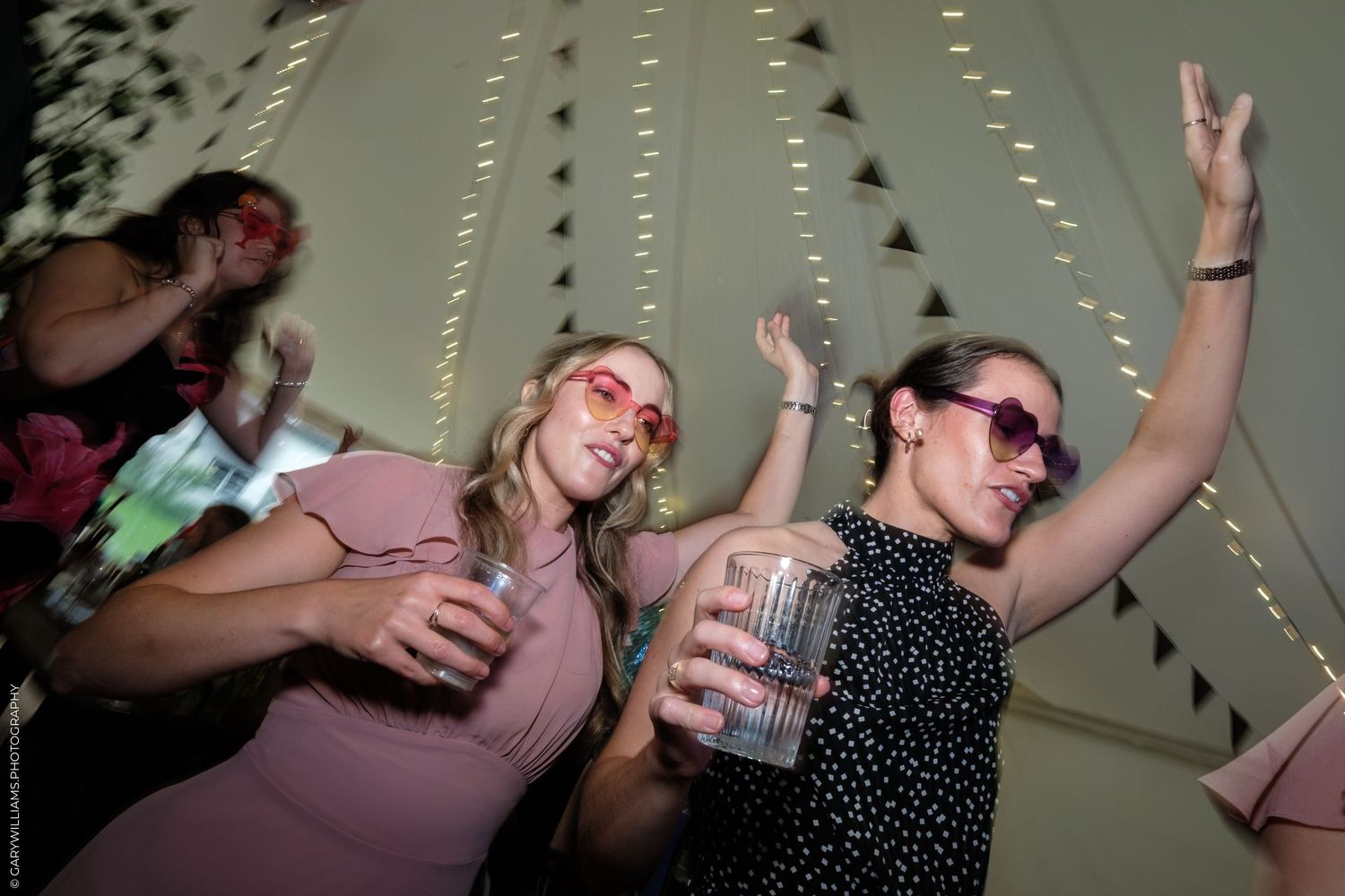 Two women dancing and holding drinks at a party inside a decorated tent with string lights.