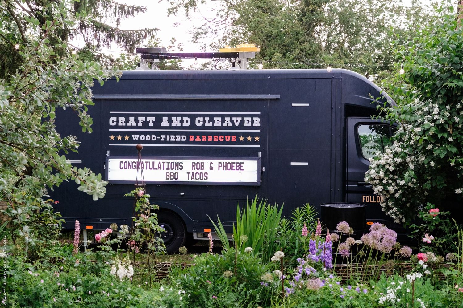 Craft and Cleaver wood-fired BBQ food truck with congratulations sign for Rob and Phoebe, surrounded by lush garden flowers.