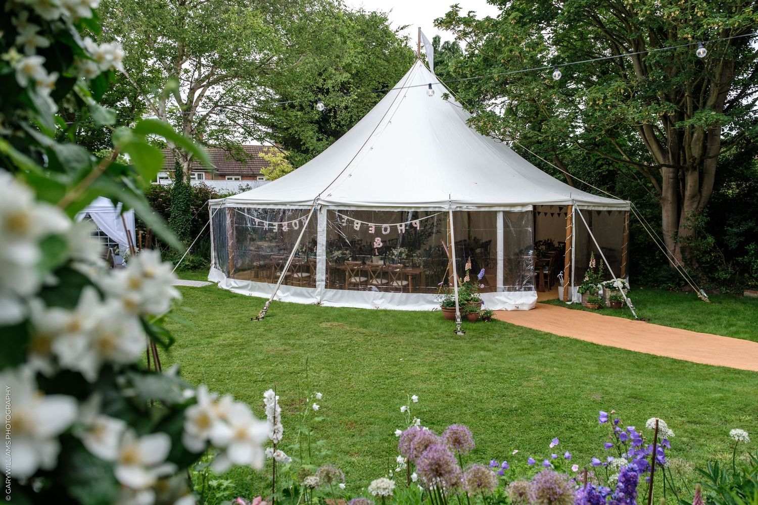 Elegant white marquee tent set in a lush garden with bunting decorations, surrounded by trees and colorful flowers.