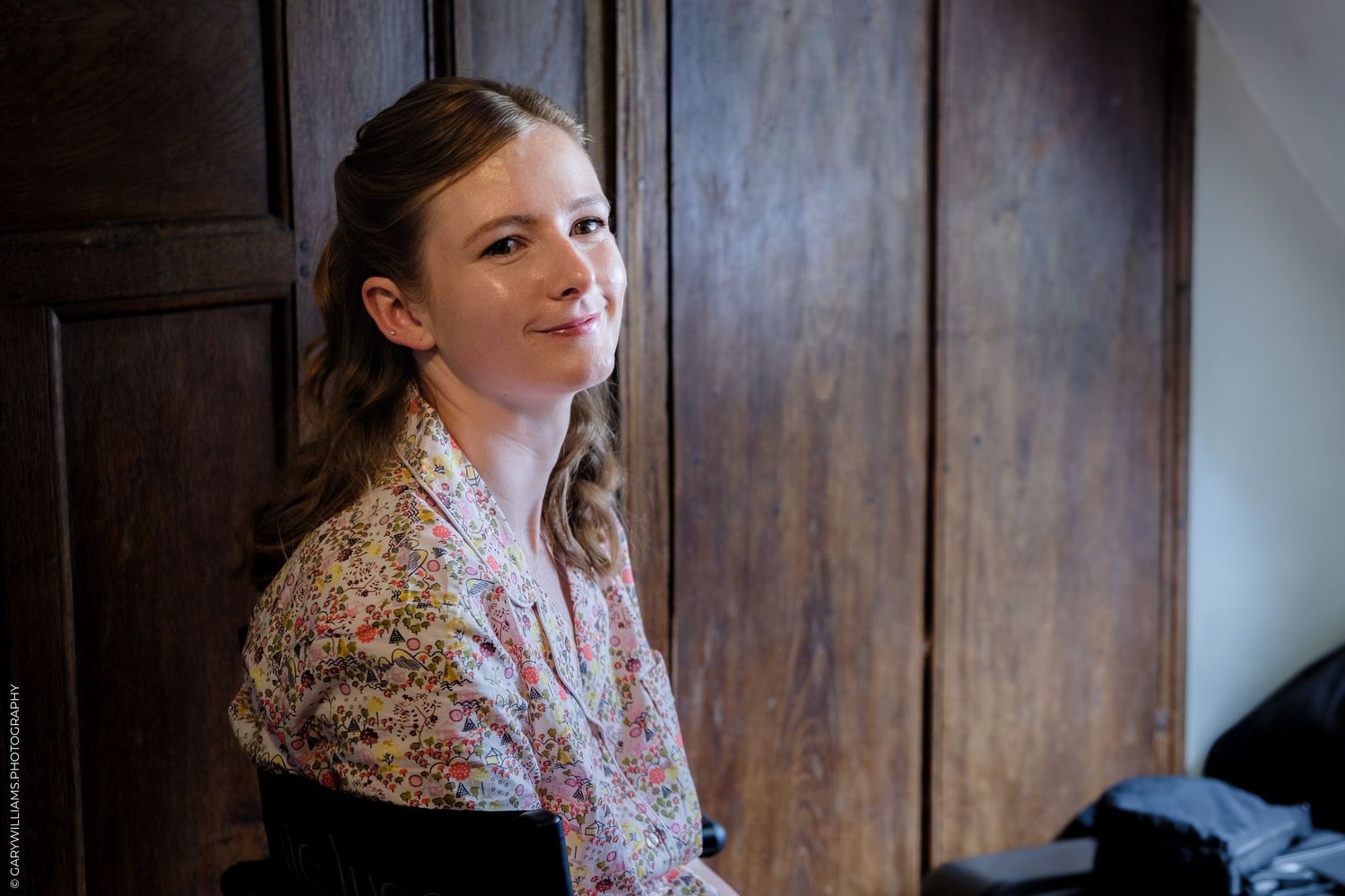 Young woman in floral blouse smiling over her shoulder, leaning against dark wooden cabinet in soft natural light.