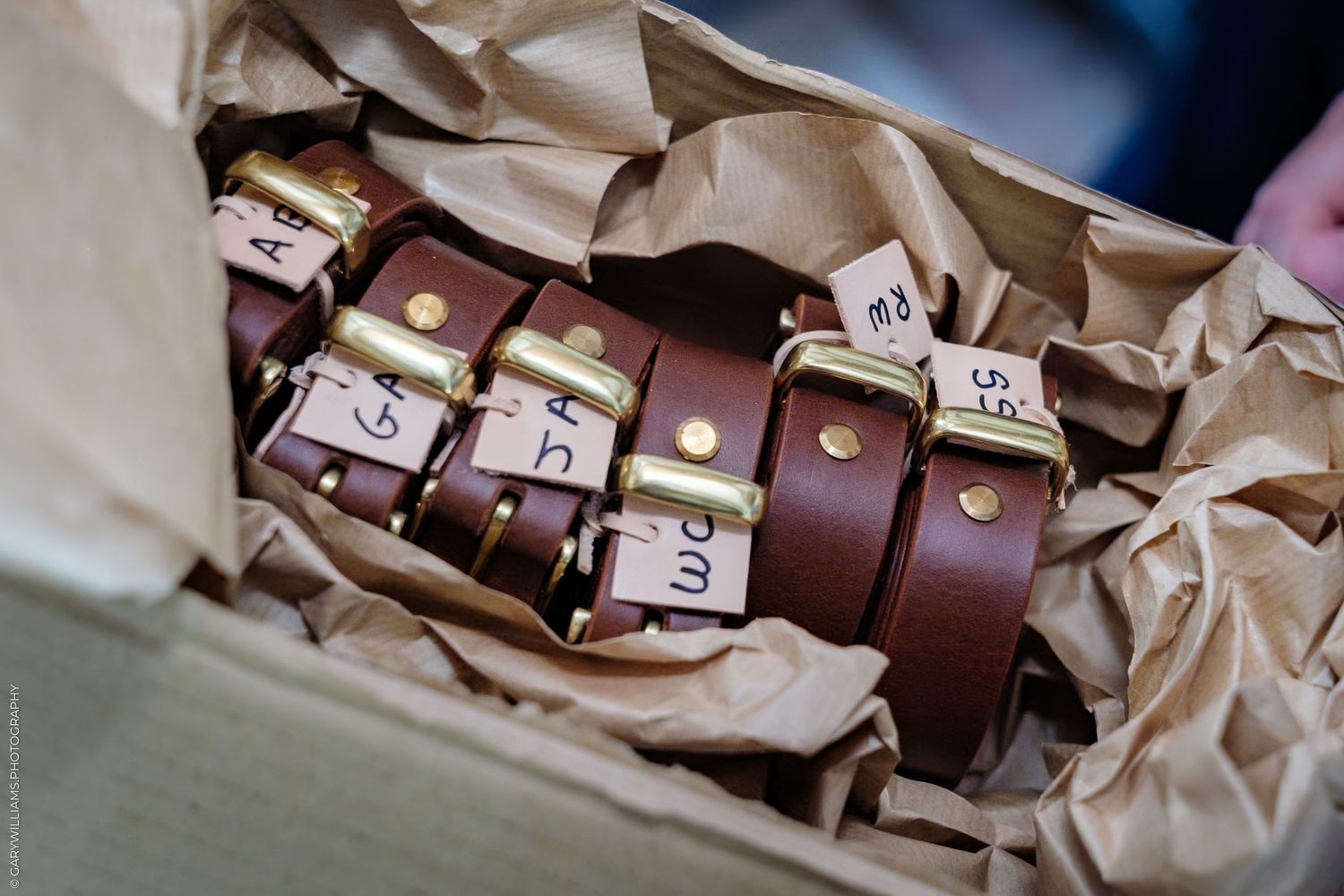Brown leather belts with gold buckles and name tags packed in a cardboard box with tissue paper.