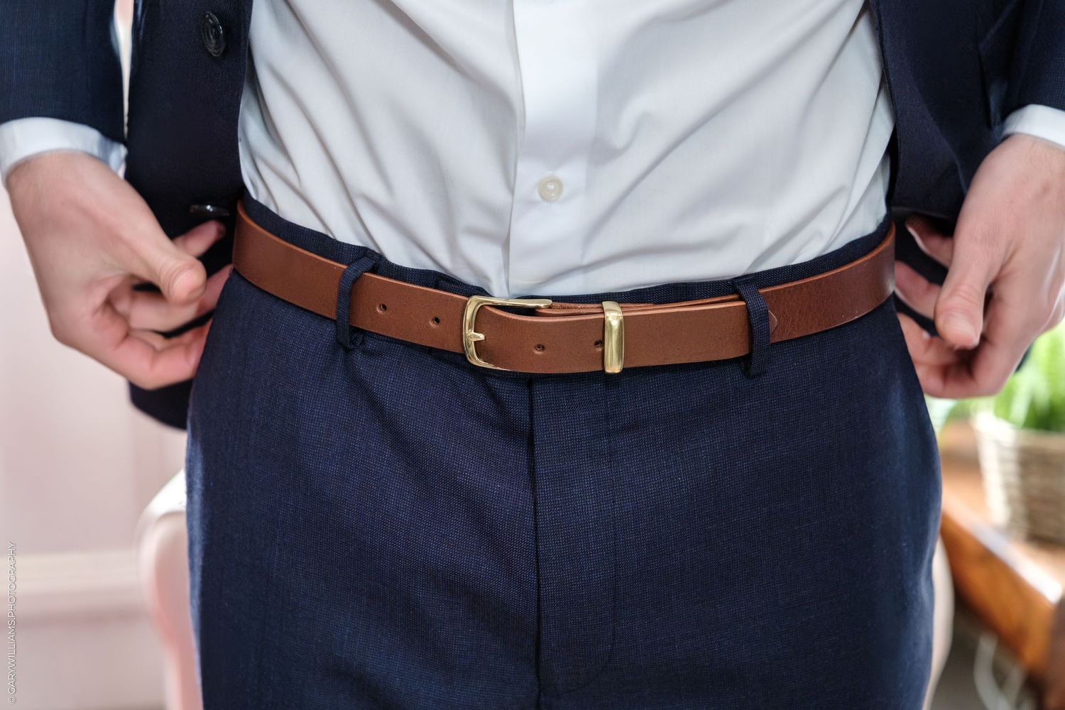 Man adjusting navy blue dress pants with a brown leather belt featuring a gold buckle, paired with a white dress shirt.