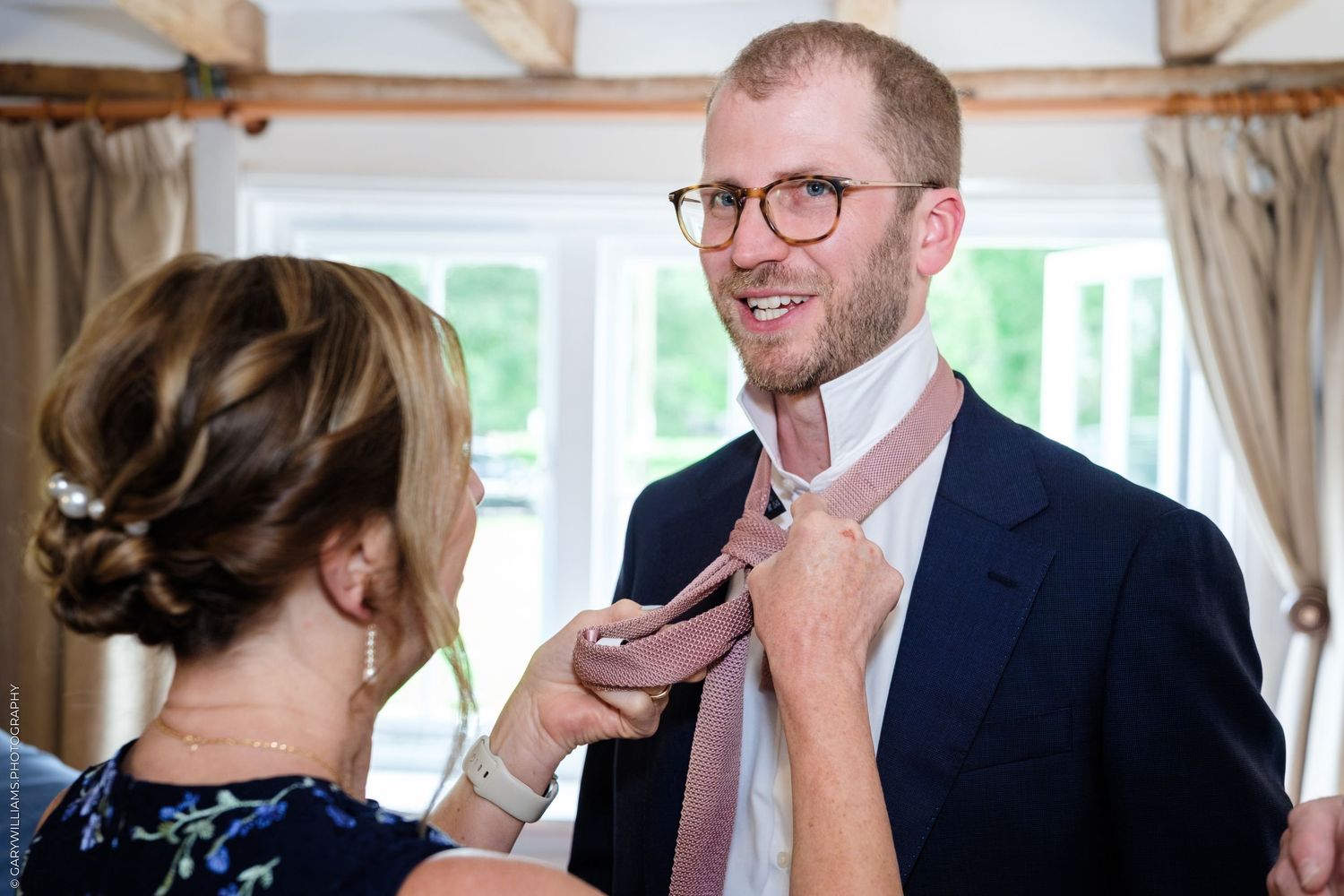 Woman adjusting pink tie on smiling groom in glasses and navy suit before wedding ceremony.