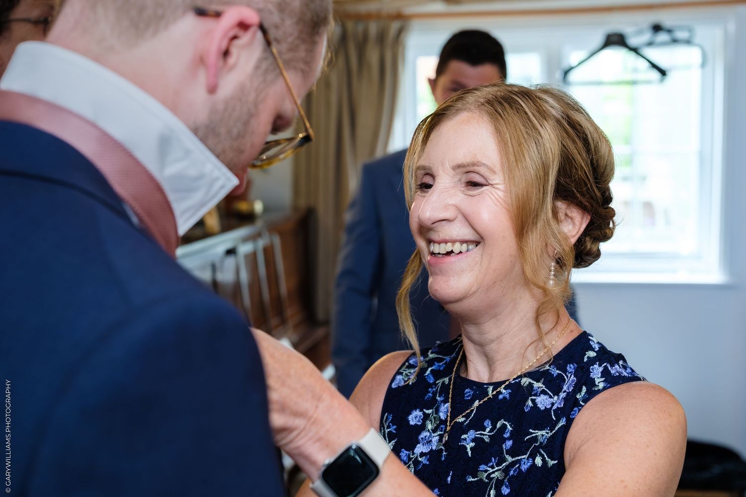 A smiling woman in a floral dress adjusts a mans suit jacket at a wedding celebration indoors.