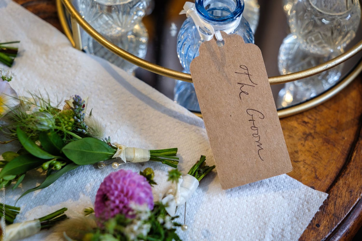 Rustic wedding place card reading The Groom on kraft paper tag beside wildflower bouquet and blue glassware.