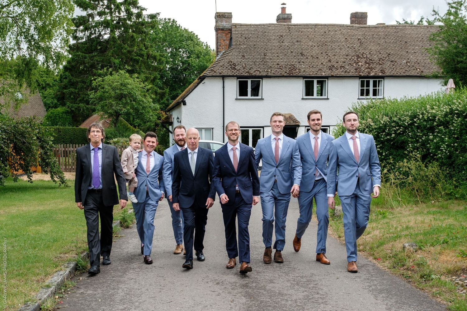 Group of groomsmen in light blue suits walking confidently down a garden path outside a countryside wedding venue.