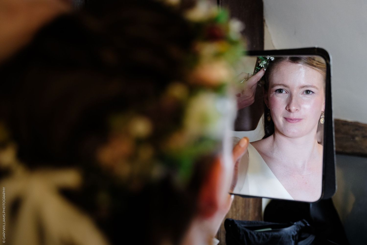 Bride checking her floral hair accessory in a handheld mirror, wearing elegant earrings and a white gown.