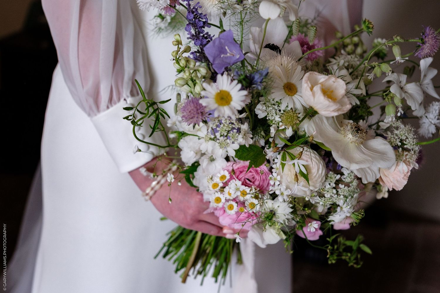Bride holding a lush wildflower wedding bouquet with daisies, lavender, and roses in soft pink and white tones.