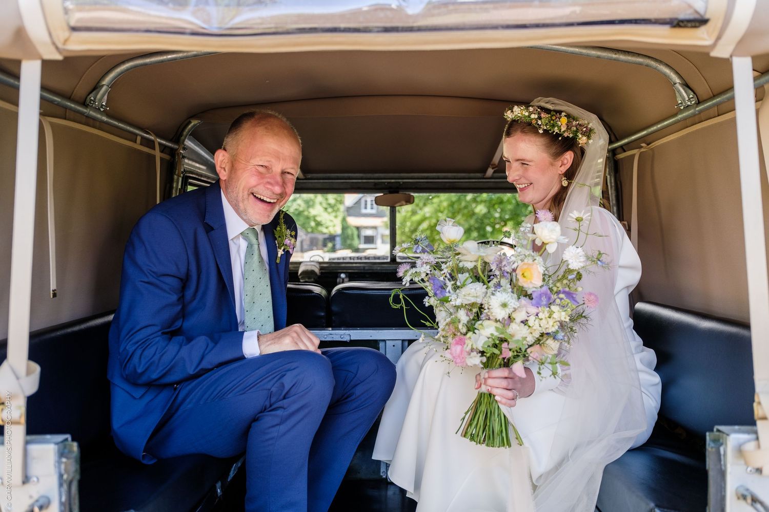 Happy bride and groom sitting inside a vintage wedding car, bride holding a pastel wildflower bouquet.