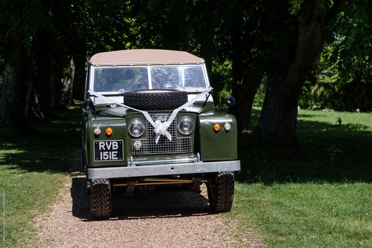 Vintage green Land Rover Series II decorated with white ribbon bow parked on gravel path surrounded by trees.