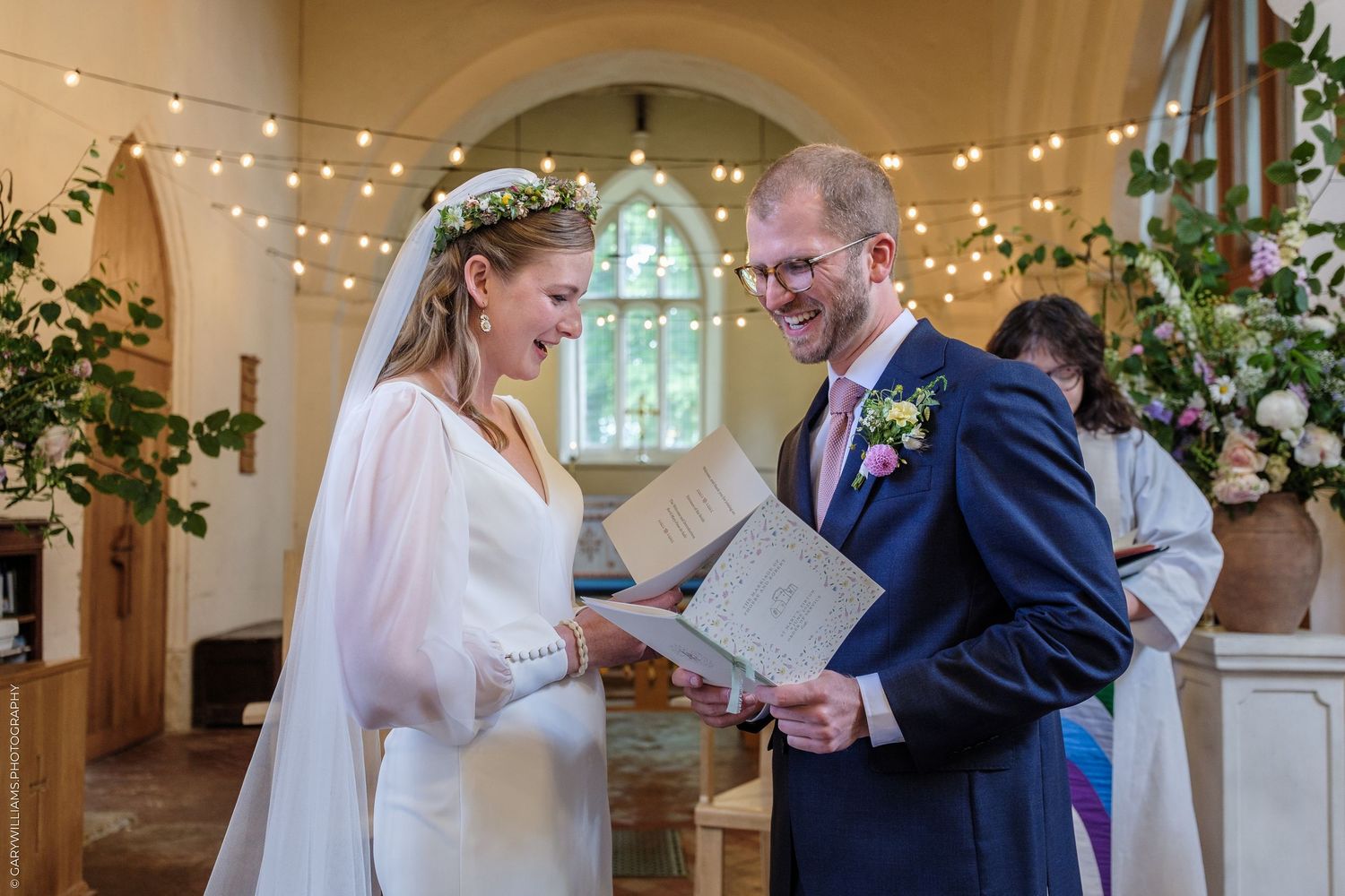 Bride and groom laughing together during church wedding ceremony, bride wearing floral crown and white gown.