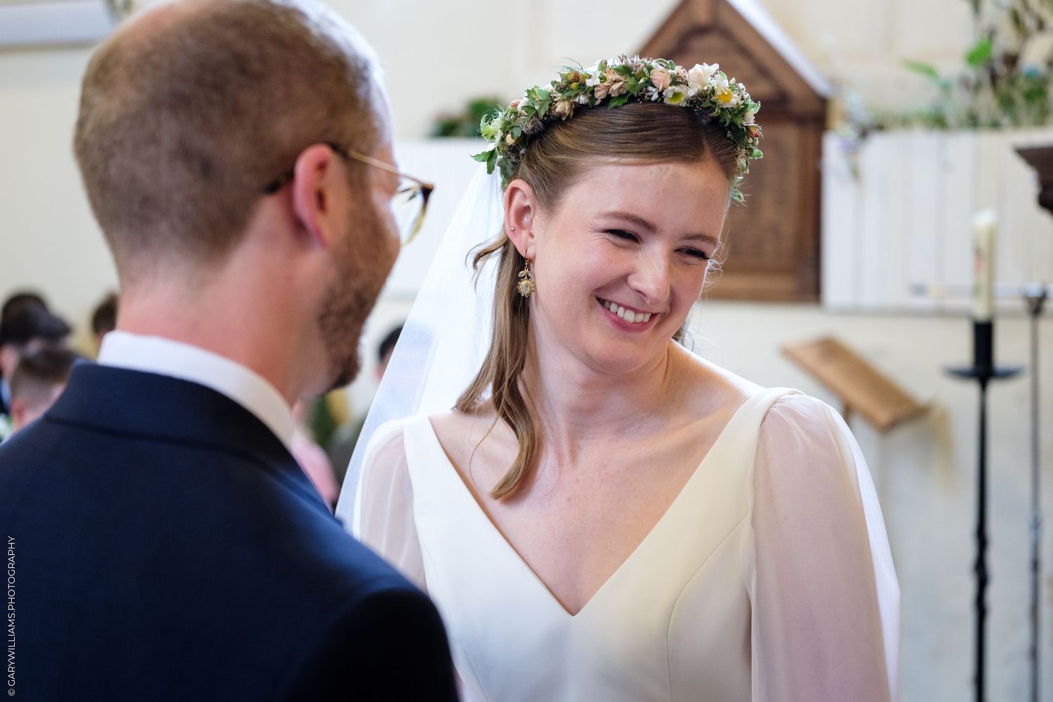 Bride in white dress and floral crown smiling at groom during wedding ceremony in church.