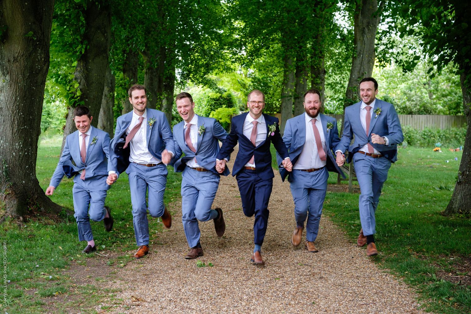 Six groomsmen in blue suits and pink ties running joyfully down a tree-lined path at a wedding.
