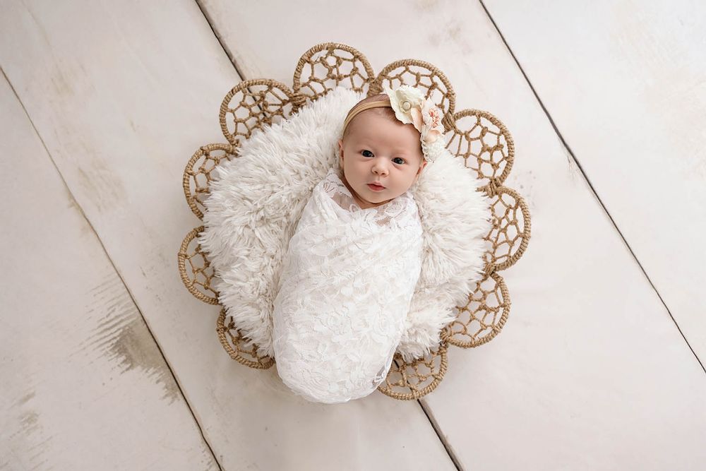 Newborn baby swaddled in white wrap with floral headband, nestled in wicker basket with fluffy fur, overhead photo.