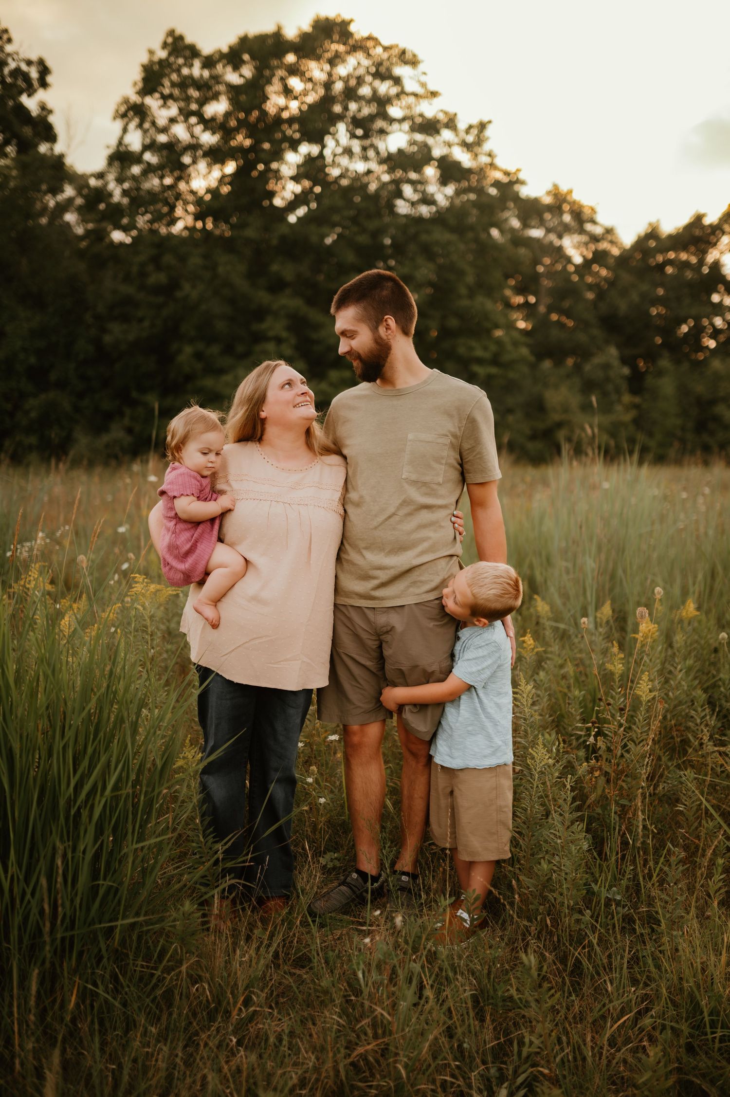 Family of four posing in a golden meadow at sunset, parents holding young children surrounded by tall grass and trees.